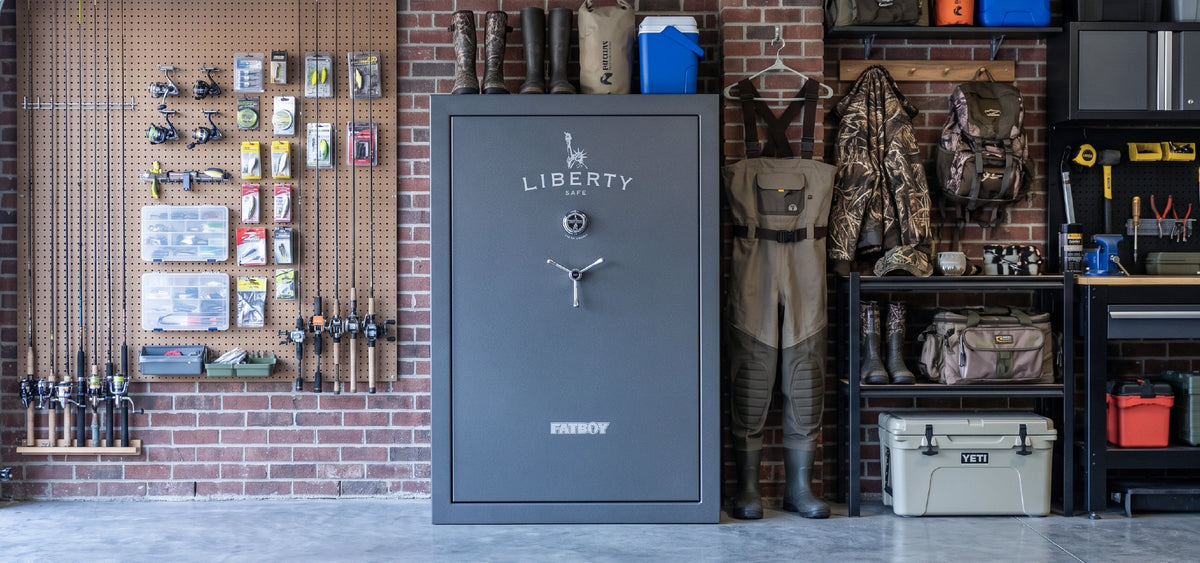 A large gray Liberty brand safe stands in a rustic workshop or garage setting with a brick wall, fishing rods on pegboards, waders, boots, and shelves filled with gear.