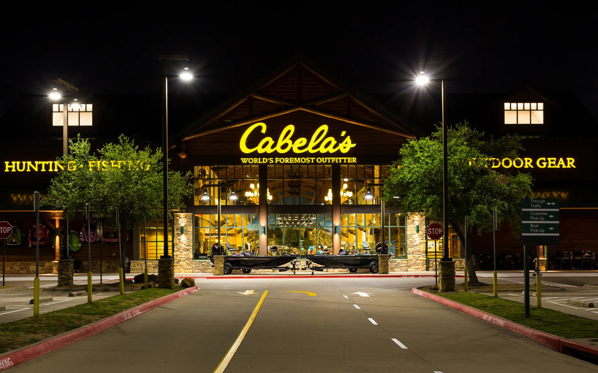 Cabela's outdoor gear storefront at night, with bright yellow signage, large glass windows, lit parking lot roads, and trees on both sides.