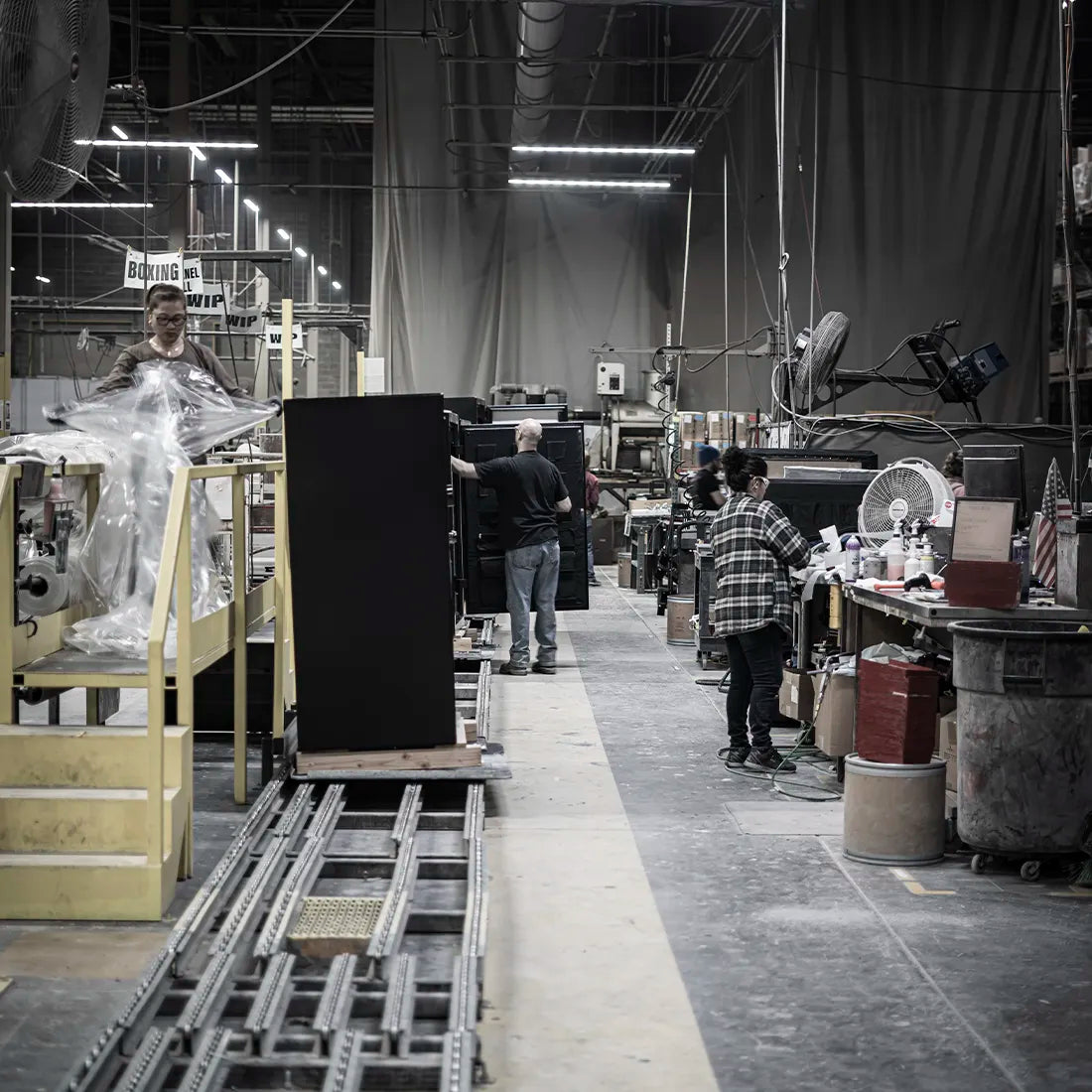A wide interior shot of a large manufacturing or workshop floor with several workers standing around industrial machinery and panels. The scene is in a real environment with visible ceiling lights, cables, worktables, and equipment, so this is Lifestyle rather than Product/Packaging.