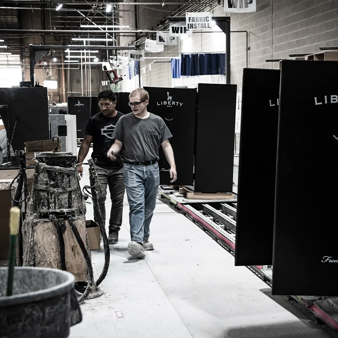 Two men walking through a workshop or fabric showroom aisle, surrounded by largeLiberty safes with white 'LIBERTY' text. The scene is indoors with industrial ceiling fixtures, tracks, and workbench equipment visible.