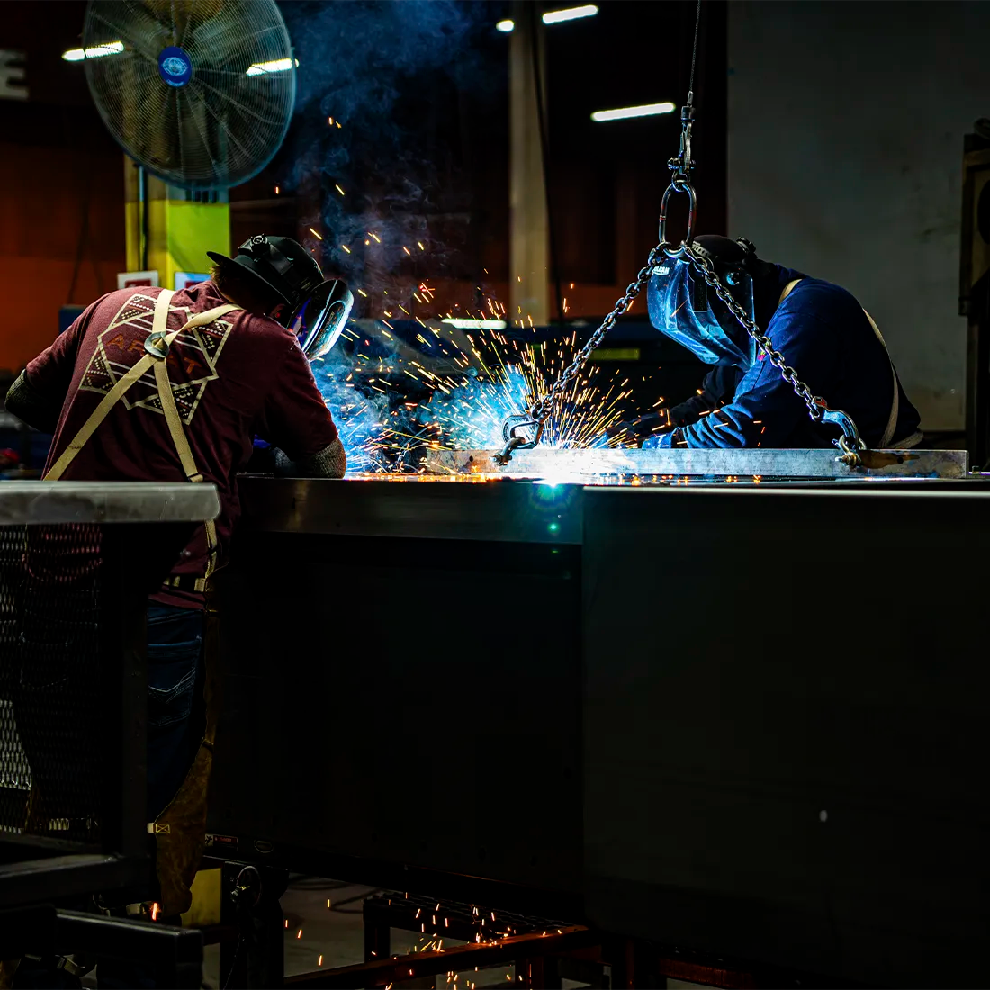 Two welders in protective gear and helmets work at a metal fabrication table, with bright blue welding light and sparks flying in a workshop.